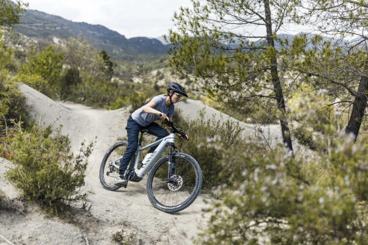 A person riding a mountain bike on a dirt trail, leaning into a turn surrounded by greenery and trees. The landscape features rolling hills in the background under a cloudy sky. The rider is wearing a helmet and casual outdoor clothing, focused on navigating the terrain.