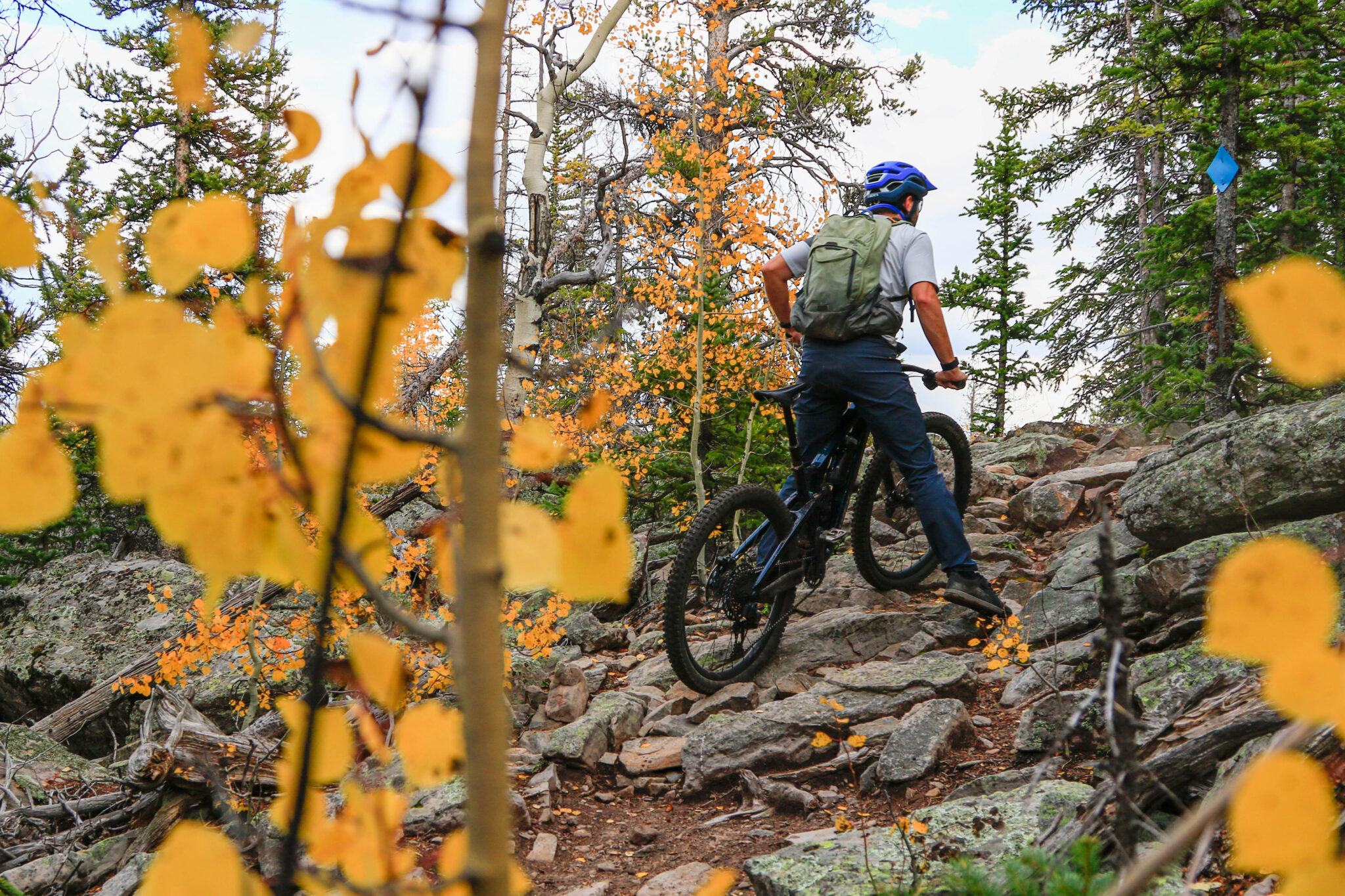 A person riding a mountain bike navigates a rocky trail surrounded by trees with yellow and orange leaves, indicating autumn. The cyclist wears a blue helmet and a backpack, focusing on the path ahead as they climb uphill through the natural landscape.