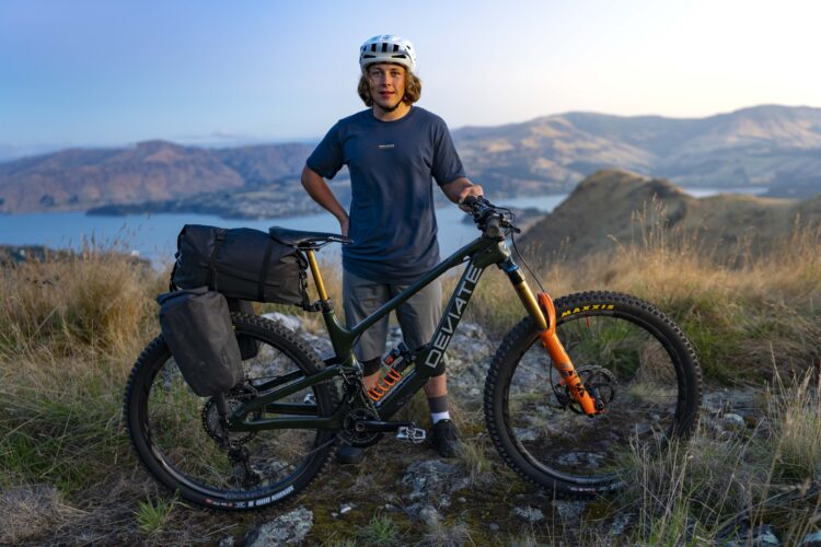 A young person stands next to a mountain bike on a grassy hillside, with a scenic view of mountains and a lake in the background. The person is wearing a helmet and a dark t-shirt, and the bike is equipped with a backpack for biking adventures. The scene is lit by warm sunlight, creating a serene outdoor atmosphere.