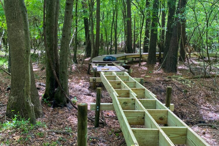 A wooden boardwalk under construction in a dense forest, surrounded by tall trees and greenery. The structure is supported by wooden posts and features a long, rectangular frame leading towards a curved section in the background. Fallen leaves and forest floor debris are visible around the area.