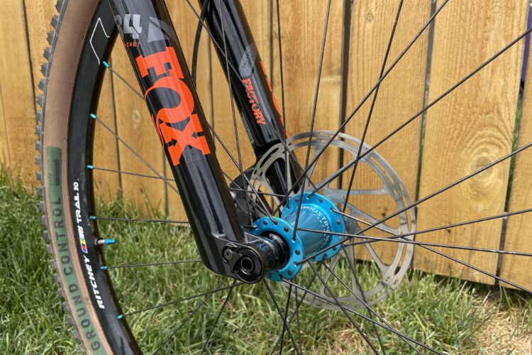 Close-up view of a mountain bike's front wheel and suspension fork, featuring a black FOX fork with orange branding, a blue hub, and a silver disc brake rotor. The background includes green grass and a wooden fence, emphasizing the outdoor setting.