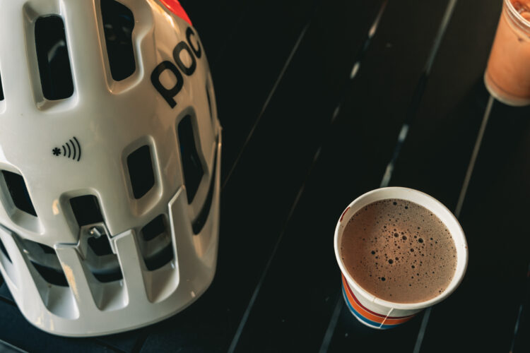 Close-up of a white bicycle helmet with ventilation holes, featuring branding, alongside a striped paper cup filled with brown liquid, placed on a dark wooden surface. The image captures a casual, outdoor setting, suggesting a blend of cycling and relaxation.