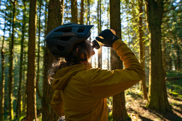 A person wearing a helmet and a yellow jacket drinks from a water bottle while surrounded by tall trees in a sunlit forest, with warm light streaming through the foliage.