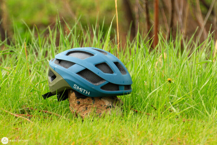 A blue cycling helmet resting on a rock in a grassy area, surrounded by fresh green grass with a hint of sunlight shining through.