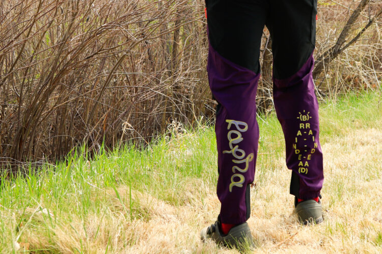 A person stands in a grassy area wearing purple and black pants, featuring a decorative design on the side. The background includes tall, dry brush and green grass. The pants are adorned with stylized text and graphics.