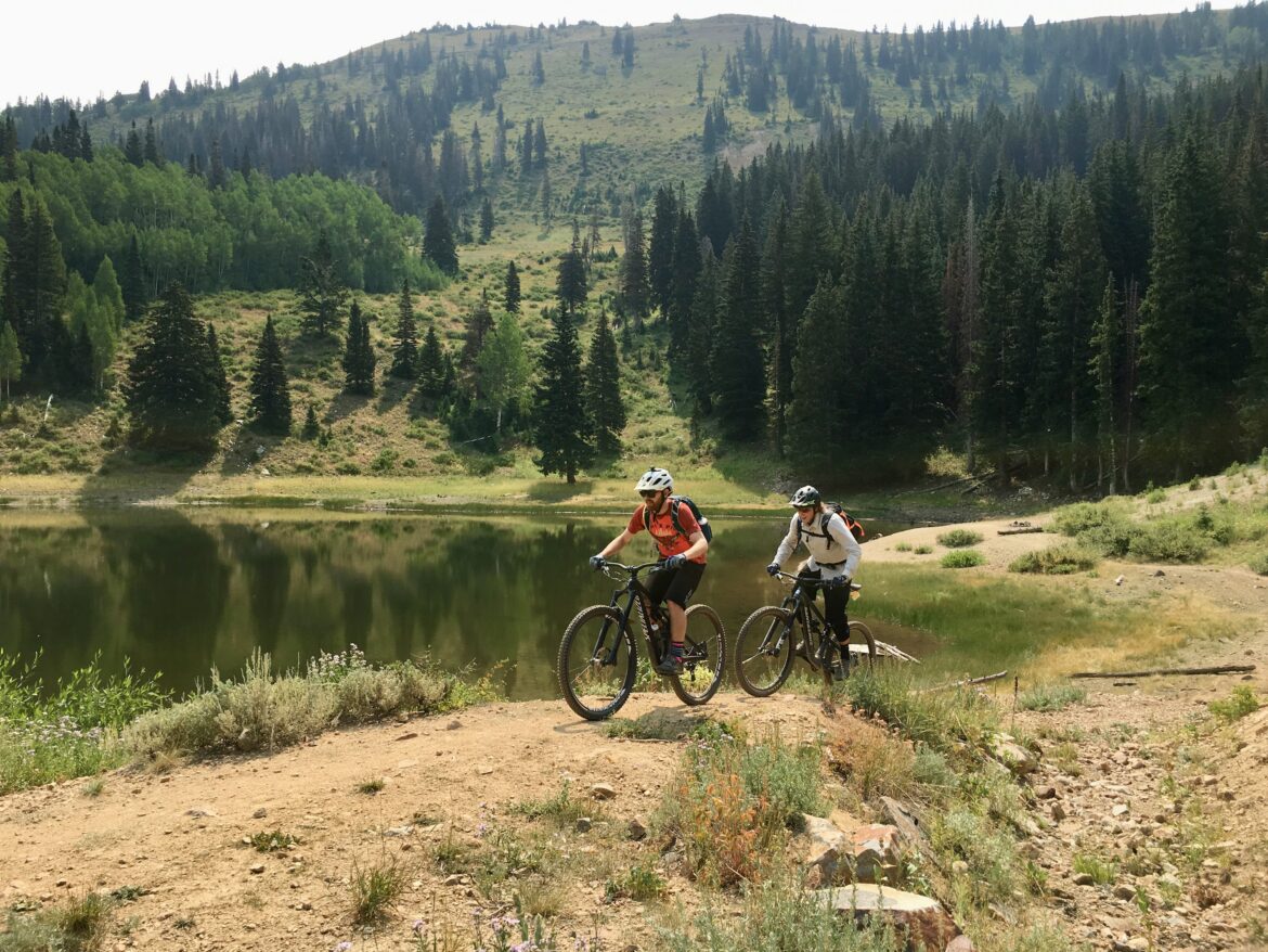 Two mountain bikers ride along a dirt path beside a calm lake, with lush green trees and a hillside in the background. The first biker is wearing an orange shirt and helmet, while the second is dressed in white. The scene captures a vibrant outdoor adventure in a beautiful natural setting.