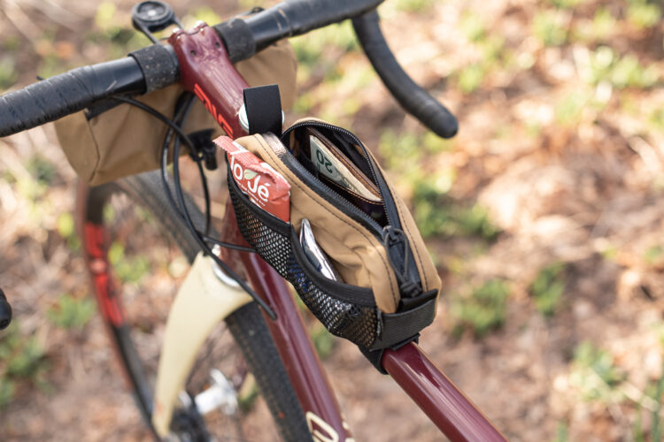 A close-up view of a maroon bicycle's handlebar featuring a brown bag attached to the frame, which contains snacks and small items. The background shows a blurred natural setting with green vegetation and soft earthy tones.