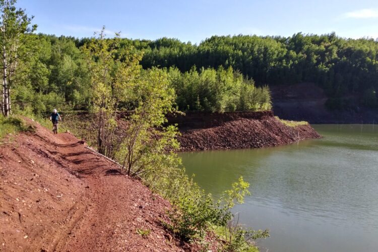 A cyclist rides along a dirt trail next to a still green lake, surrounded by lush green trees and a blue sky. The trail is slightly elevated, with a rocky terrain and vibrant foliage on either side.
