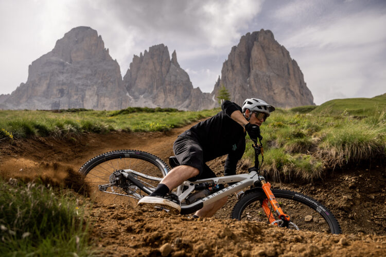 A cyclist wearing a helmet and sunglasses leans into a turn on a mountain bike, kicking up dirt on a rugged trail. Majestic mountains loom in the background under a partly cloudy sky, creating a dramatic outdoor scene.