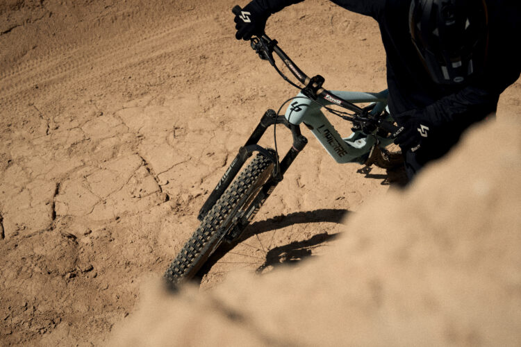 A mountain biker navigating a sandy terrain on a light blue bicycle, focused on maintaining balance and control. The rider wears a black long-sleeve shirt and gloves, with the bike's tire and distinctive frame visible in the foreground. The setting features dry, cracked earth, suggesting a challenging riding environment.