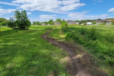 A winding dirt path through a lush green field under a partly cloudy blue sky, with trees on one side and a glimpse of buildings in the distance. Campus Life Single Track mountain bike trail.