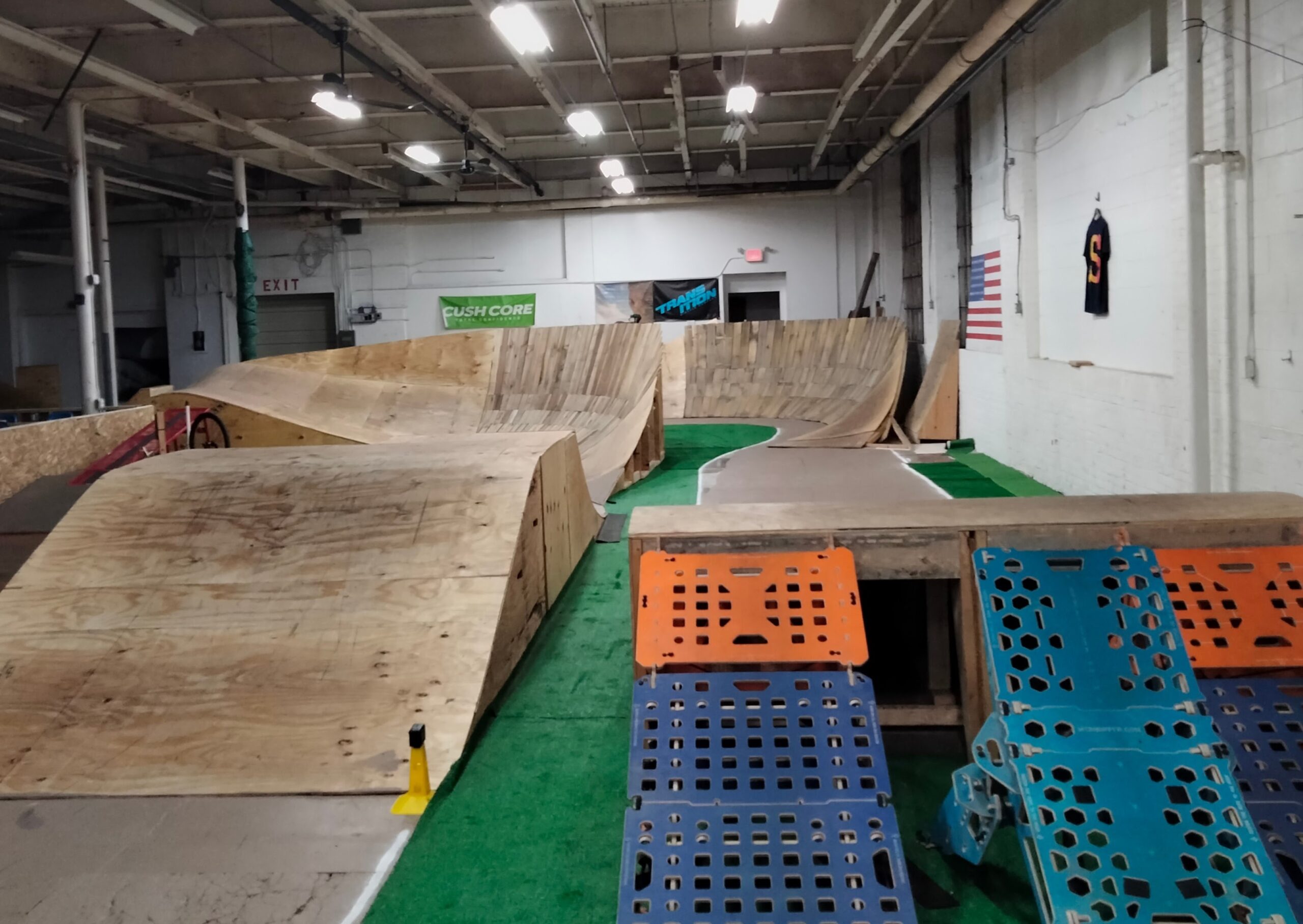 Indoor skatepark featuring wooden ramps, a green turf surface, and various obstacles including jumps and inclines. Bright overhead lights illuminate the space, which has an open layout with a wall displaying a flag and promotional banners. "Flow Track" mountain bike trail.