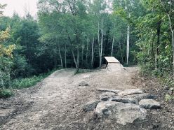 A dirt trail in a forested area, featuring a small jump ramp made of wood. Surrounding the trail are lush green trees and rocky terrain. Double Dragon mountain bike trail.