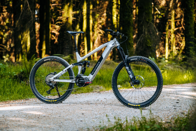 A modern mountain bike positioned on a gravel path, surrounded by lush green foliage and tall trees. The bike features a sleek silver and black design, showcasing its frame, large tires, and high-quality components, with an emphasis on trail-ready capabilities. Sunlight filters through the forest, highlighting the natural beauty of the setting.