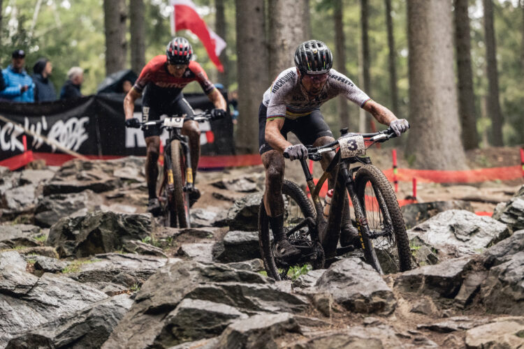 A mountain bike race featuring two cyclists navigating a rocky terrain in a forested area. One rider is in the foreground, covered in mud and wearing a white jersey with black and colorful accents, while the second rider trails behind in a red jersey. Spectators can be seen in the background, and a banner is partially visible amidst the trees. The scene captures a dynamic moment in the race, emphasizing the challenging conditions.