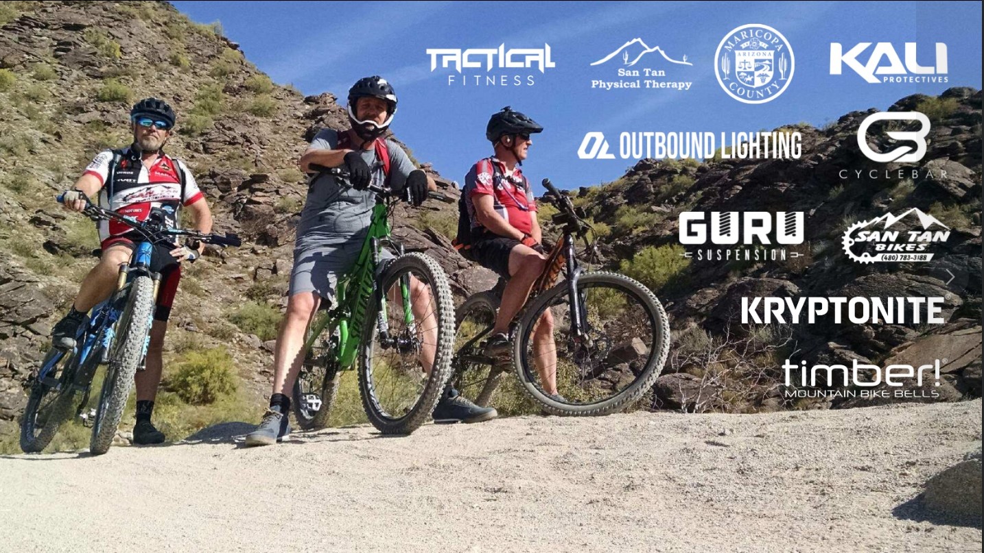 Three mountain bikers pose on a rocky trail, showcasing their bikes. The landscape features rugged terrain and sparse vegetation under a clear blue sky. Various brand logos related to biking and fitness are displayed in the background, including Tactical Fitness, San Tan Physical Therapy, and Kali Protectives.