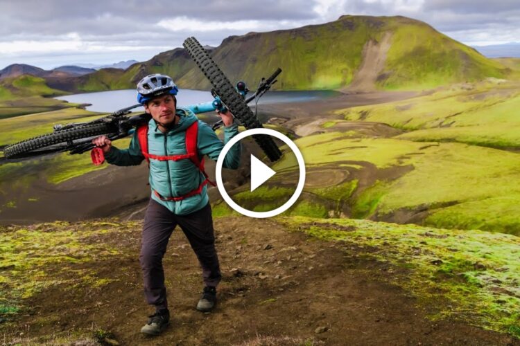 A person carrying a mountain bike on their shoulder while hiking on a green hillside, with a scenic landscape featuring mountains and a body of water in the background. The individual is wearing a helmet and outdoor gear, with a focus on adventure and exploration.
