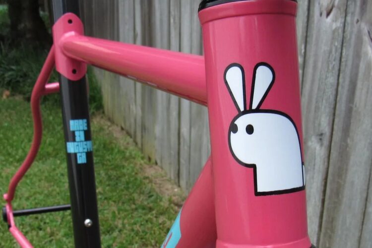 Alt text: Close-up of a vibrant pink bicycle frame featuring a playful white rabbit sticker on the head tube, with a blurred wooden fence and grass in the background.