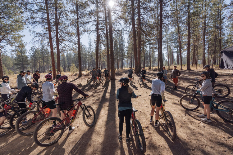 A group of people gathered in a forest setting, having a discussion while standing next to their mountain bikes. The scene is surrounded by tall trees and dappled sunlight. Some participants are wearing helmets, and the setting appears to be suitable for outdoor biking activities.