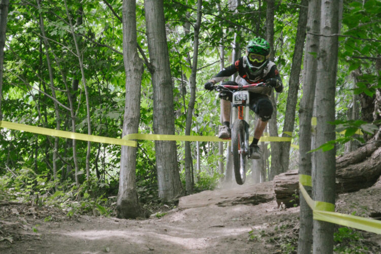 A mountain biker in protective gear jumps over a dirt path while navigating a forested trail marked with yellow tape. Dust is kicked up from the ground as the biker catches air above the trail, surrounded by greenery and trees.