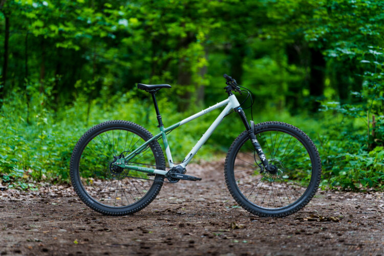 A mountain bike with a white and green frame is parked on a dirt trail surrounded by lush green foliage. The setting suggests a serene, natural environment, perfect for outdoor cycling adventures.
