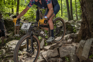 A mountain biker wearing a colorful jersey and gloves navigates a rocky trail in a dense forest, captured mid-jump with a focused expression. The bike is in an upward position as the rider skillfully maneuvers over the uneven terrain, with green trees surrounding the scene and spectators visible in the background. Oak Mountain State Park Bump Trail mountain bike trail.