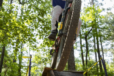 A mountain biker in protective gear jumps off a wooden ramp surrounded by trees, capturing a moment of adrenaline and skill during an outdoor cycling event. The biker is dressed in a colorful shirt and wears a helmet with goggles, emphasizing the action in a forested setting. Oak Mountain State Park Bump Trail mountain bike trail.