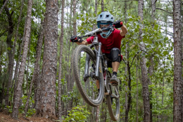 A mountain biker wearing a helmet and protective gear jumps off a dirt ramp in a forested area. The biker is airborne, with trees and lush greenery surrounding the trail. Oak Mountain State Park mountain bike trail.