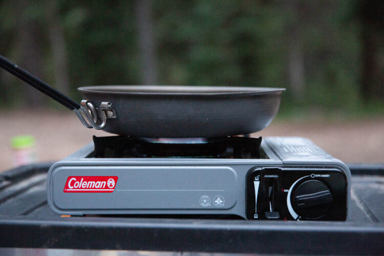 A portable Coleman camping stove with a black frying pan on top, set against a blurred outdoor background. The stove features a control knob and a brand logo.