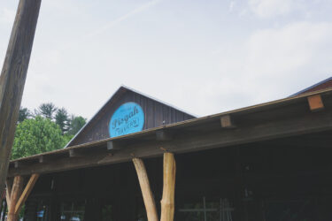 Image of the exterior of Pisgah Tavern, featuring a wooden structure with a blue sign that reads "Pisgah Tavern." The background includes trees and a partly cloudy sky, with wooden support beams visible in the foreground.