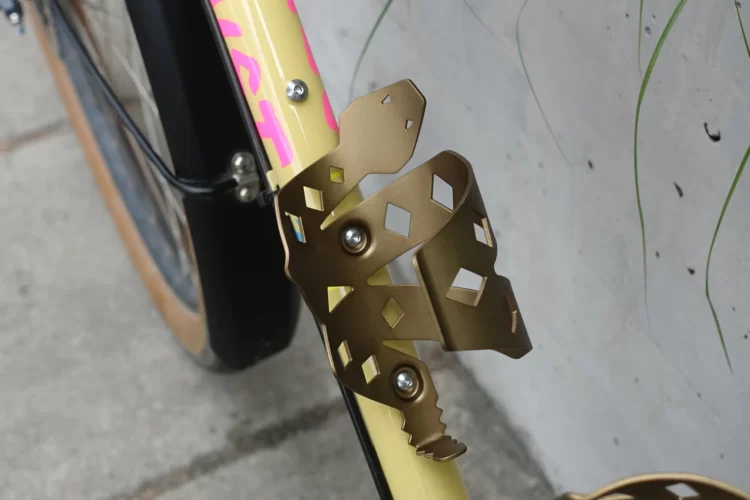 A close-up view of a gold bike water bottle cage attached to a yellow bicycle frame. The cage features a unique cut-out design and is mounted near the front of the bike, showcasing a colorful branding element in pink. The background consists of a textured concrete surface and some greenery.