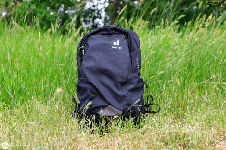 A black backpack sitting on grass in a natural outdoor setting. The backpack features the logo of the brand "Deuter" prominently displayed on the front. Surrounding the backpack is tall, green grass, indicating a summery or springtime environment.