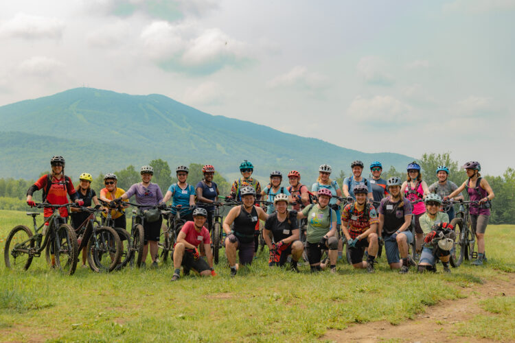 A group of 30 mountain bikers posing outdoors, wearing helmets and cycling gear, with several mountain bikes in the foreground. The backdrop features a lush green landscape with rolling hills and a distant mountain under a partly cloudy sky.