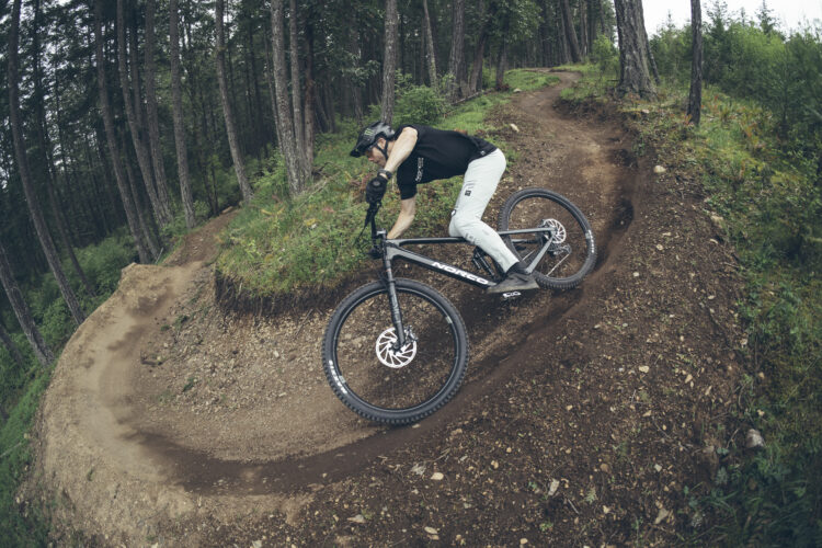 A mountain biker in a black shirt and light gray pants leans into a curved dirt trail surrounded by tall trees. The biker rides a black Norco bicycle, navigating the trail on a cloudy day.