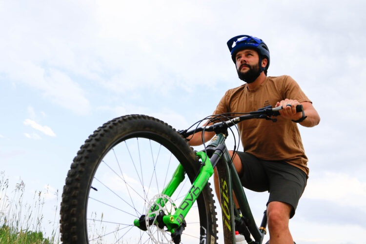 A man wearing a blue helmet and a brown shirt rides a green mountain bike on a trail. He is looking forward with determination, and the sky is partly cloudy in the background. Tall grass is visible on either side of the trail.