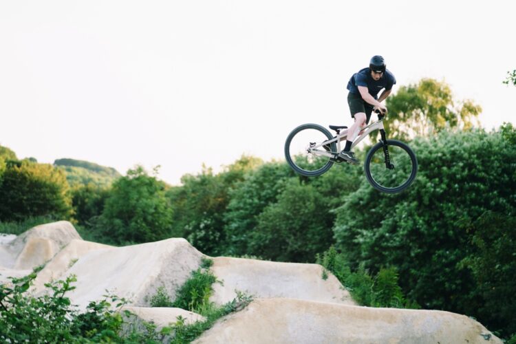A person riding a mountain bike is performing a jump over dirt mounds in a green outdoor setting, showcasing a dynamic biking maneuver. The scene captures the rider mid-air against a backdrop of trees and a clear sky.