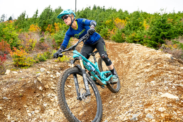 A mountain biker navigates a dirt trail surrounded by lush greenery and vibrant fall foliage, wearing protective gear and a blue jersey, while leaning into a turn on their turquoise bike.