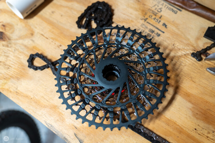 A close-up view of a black bicycle cog assembly placed on a wooden workbench, surrounded by bicycle chain links and tools. The cog features an intricate design with openings that give it a lightweight appearance, highlighting its mechanical details.