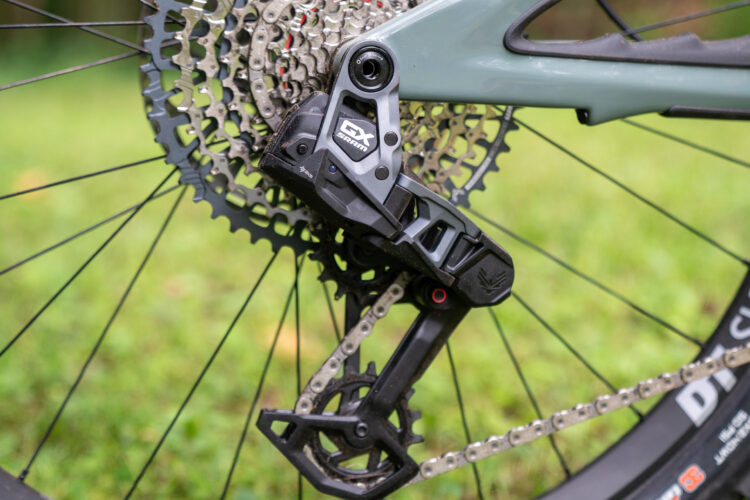 Close-up of a mountain bike rear derailleur mounted on a cassette and chain, set against a blurred green background of grass and trees. The derailleur is black with a silver label, and the cassette features a series of interlocking metallic gears. The bike's tire is partially visible in the corner.