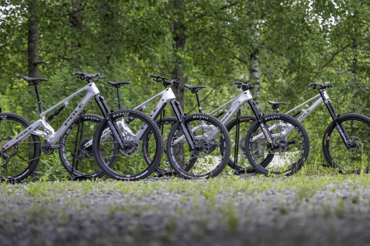 A row of four mountain bikes in a natural outdoor setting, featuring a mix of silver and black frames with prominent branding. The bikes are positioned on gravel ground, surrounded by green foliage and trees, suggesting an adventurous atmosphere.