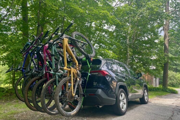 A black SUV parked on a gravel road with multiple mountain bikes attached to a hitch-mounted bike rack. The bikes, in various colors including yellow, green, and purple, are secured upright and have their wheels resting on the ground. Surrounding the vehicle are lush green trees, creating a natural outdoor setting.