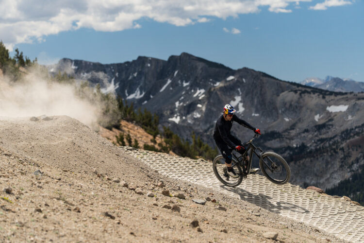 A mountain biker performs a trick on a dirt trail surrounded by mountains, with clouds in the sky and dust rising from the ground. The cyclist wears a helmet and riding gear and is positioned on a textured surface designed for biking.