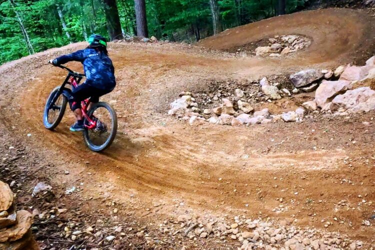 A mountain biker navigates a winding dirt trail through a lush forest, showcasing sharp turns and natural obstacles. The cyclist is dressed in activewear and wearing a helmet, emphasizing the adventurous spirit of outdoor biking. Surrounding trees provide a vibrant green backdrop.