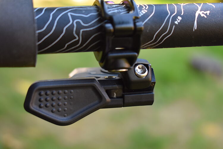 Close-up image of a bicycle handlebar featuring a black control switch attached to the side, with a textured grip for ease of use. The handlebar has a topographic design printed on its surface, and a black rubber grip is visible at one end. The background is softly blurred, suggesting an outdoor setting.