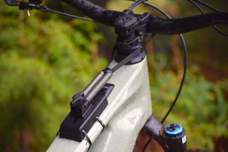 Close-up image of a mountain bike's handlebar area, featuring a matte green frame with raindrops, a black headset, and a cable routing system. The image captures the intricate details of the bike's components, showcasing a metal spring and a mount at the top. Soft green foliage is blurred in the background, indicating an outdoor setting.