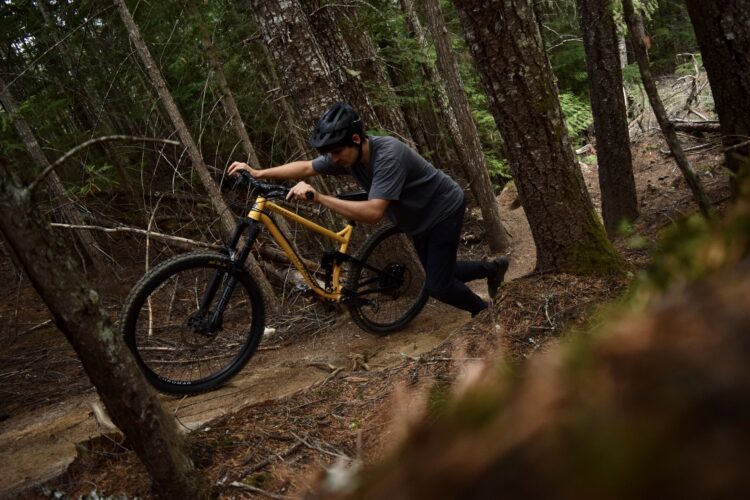 A person in a helmet pushes a yellow mountain bike up a steep trail through a dense forest. The terrain is rugged, with pine trees and fallen branches visible in the surroundings. The individual is focused on navigating the challenging path.
