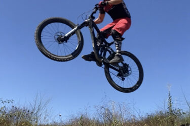 A mountain biker performing a jump over a dirt track, suspended in mid-air against a clear blue sky, surrounded by sparse vegetation. The rider is wearing a helmet and protective gear, and the bike's front wheel is elevated off the ground. San Clemente Singletracks mountain bike trail.