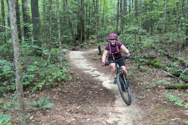 A person riding a mountain bike along a winding dirt trail in a lush green forest, wearing a pink helmet and cycling attire, with trees and underbrush surrounding the path. Rocky Ridge mountain bike trail.