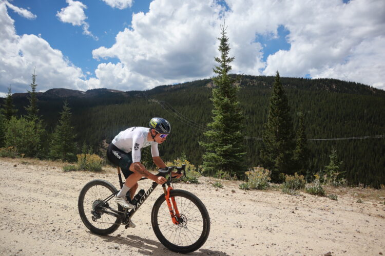 A cyclist rides on a dirt path surrounded by lush green trees and rolling hills under a partly cloudy blue sky. The cyclist is focused and wearing a helmet, sunglasses, and a racing jersey, showcasing an energetic outdoor lifestyle.