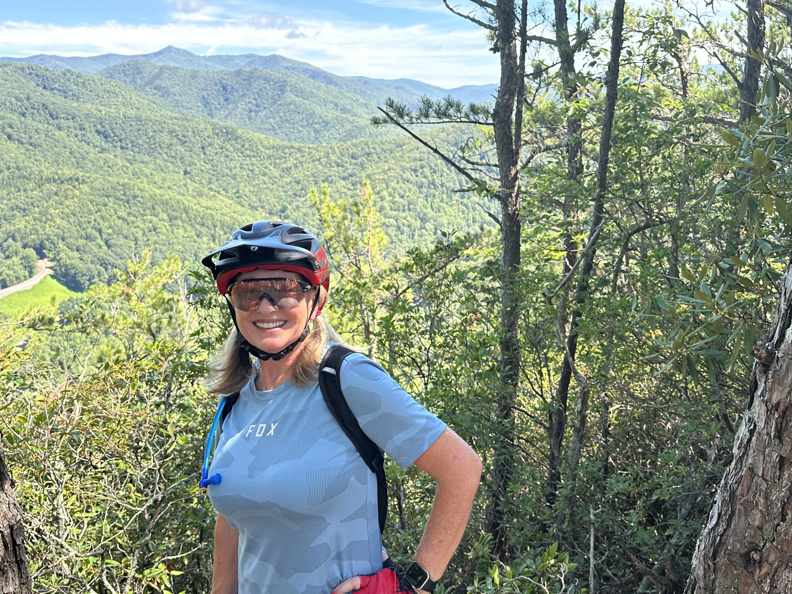A smiling woman wearing a red helmet and sunglasses stands in a wooded area, surrounded by green hills and mountains in the background. She is dressed in a light blue shirt and has a water reservoir on her back, indicating she is engaged in outdoor biking or hiking activities. Bernard Mountain Trail mountain bike trail.